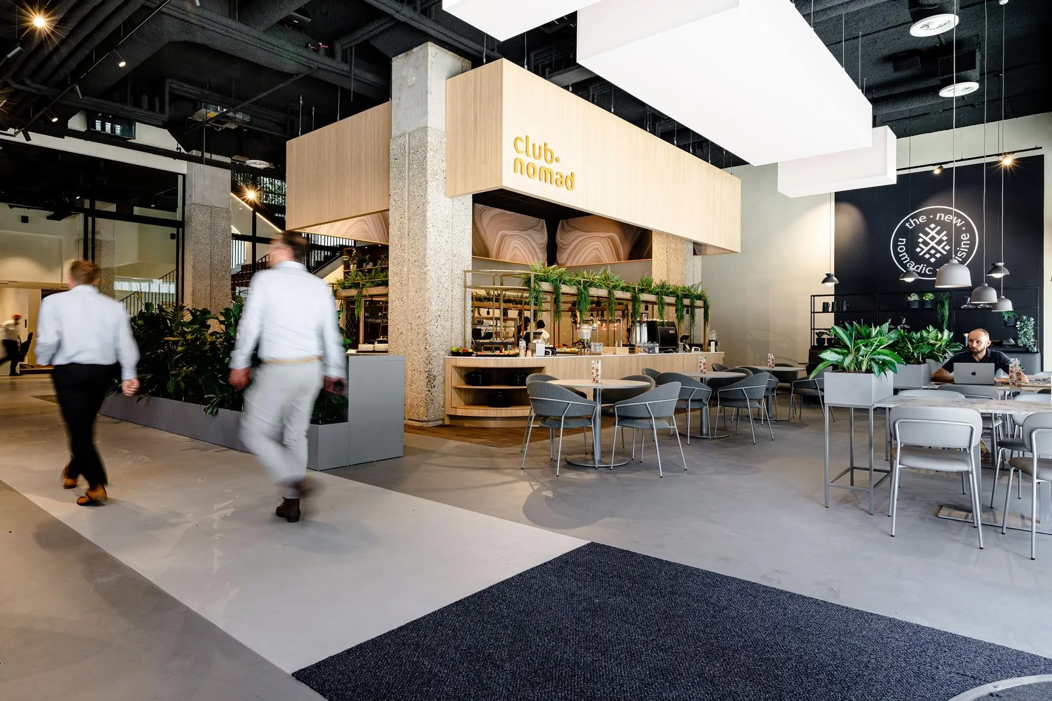 Modern ingerichte horecaruimte genaamd Club Nomad aan de Aert van Nesstraat, met zitplaatsen, groene planten en werkende mensen.